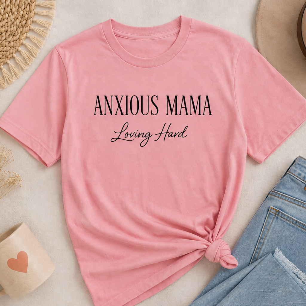 A pink short-sleeve t-shirt with the text 'ANXIOUS MAMA Loving Hard' printed in black, styled in a flat lay with light blue jeans, a straw hat, a mug, and dried grass on a light beige background.